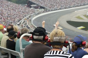 A DSLR photo of a couple of senior fans at a racing event. There is a crowd of people, the stadium is full and there is racing cars on the track. Some people are wearing headsets. There is also one person with arms raised in the air enjoying the race.